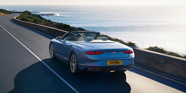 Bentley Singapore New Bentley Continental GTC Azure convertible rear view in Light Windsor blue paint with 22 inch ten spoke wheels driving dynamically on an ocean road in the sunshine