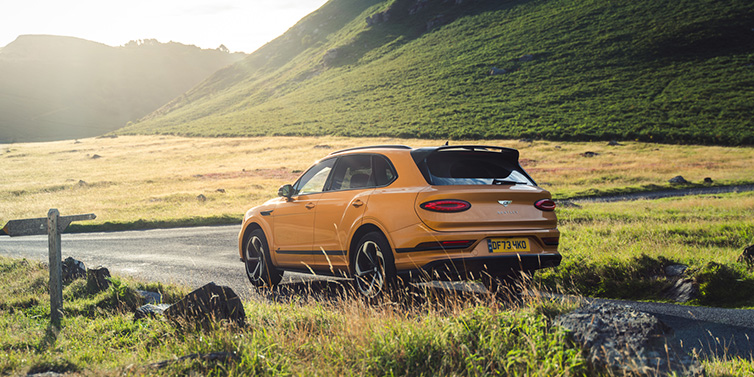 Bentley Singapore Bentley Bentayga S SUV rear three quarter in Sunburst Gold paint on a road surrounded by green hills